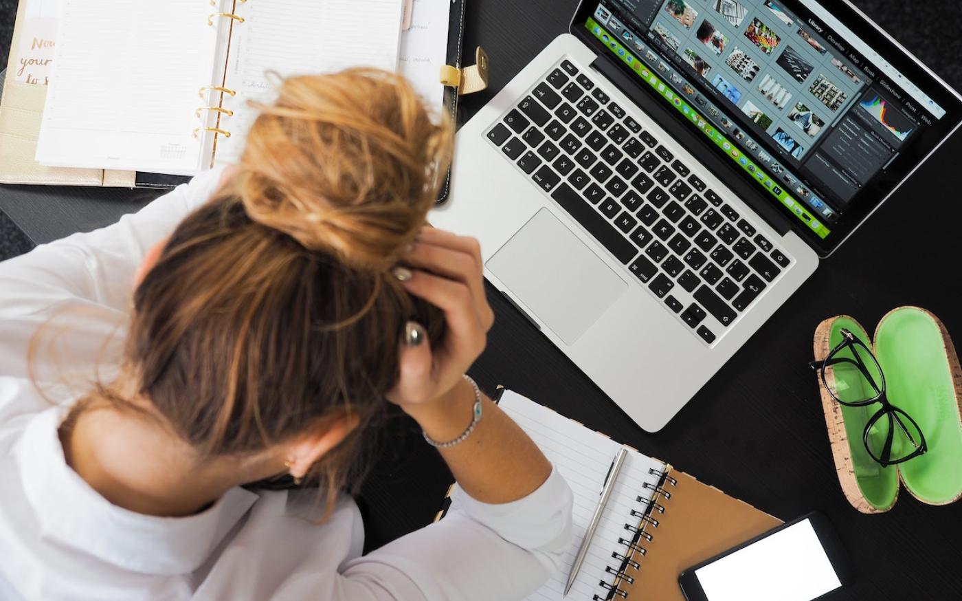 Top-down image of a woman sitting at a cluttered desk. She's holding her head and looking down.