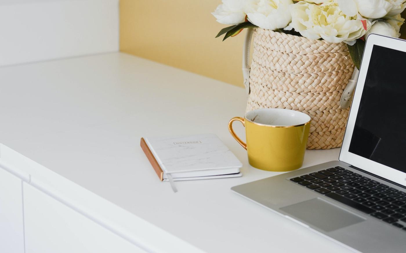 A desk with a laptop, a yellow coffee cup, and a notebook on it. Next to the laptop is a vase filled with white flowers